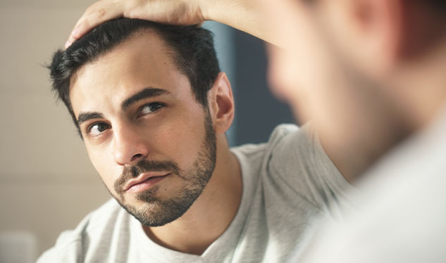 A man in a white t-shirt examines his hair in a mirror, with one hand on his head.