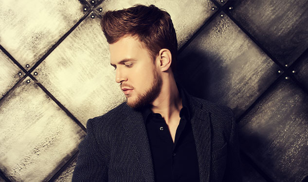 A man with styled hair and a beard looks downward against a textured gray wall, wearing a dark jacket and shirt.