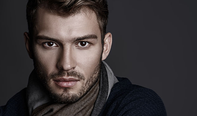Portrait of a man with short hair and a beard, wearing a scarf and dark clothing, looking directly at the camera against a dark background.