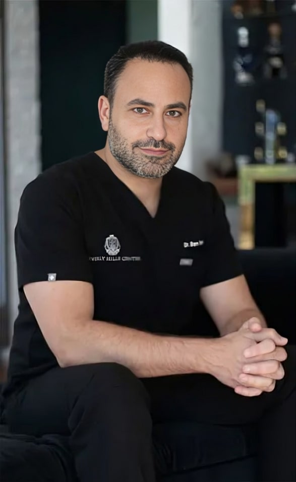 A man in black medical scrubs sits on a black chair with his hands clasped, looking at the camera.